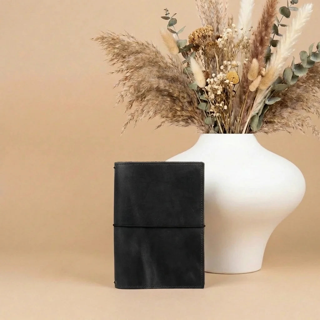 Black leather journal next to a white vase with dried flowers on a beige background