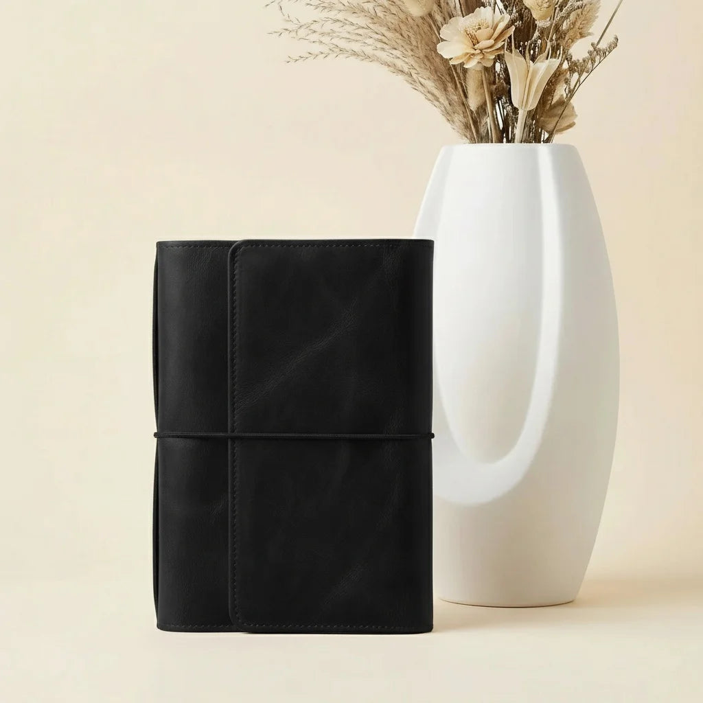 Black leather-bound notebook next to a white vase with dried flowers on a beige background