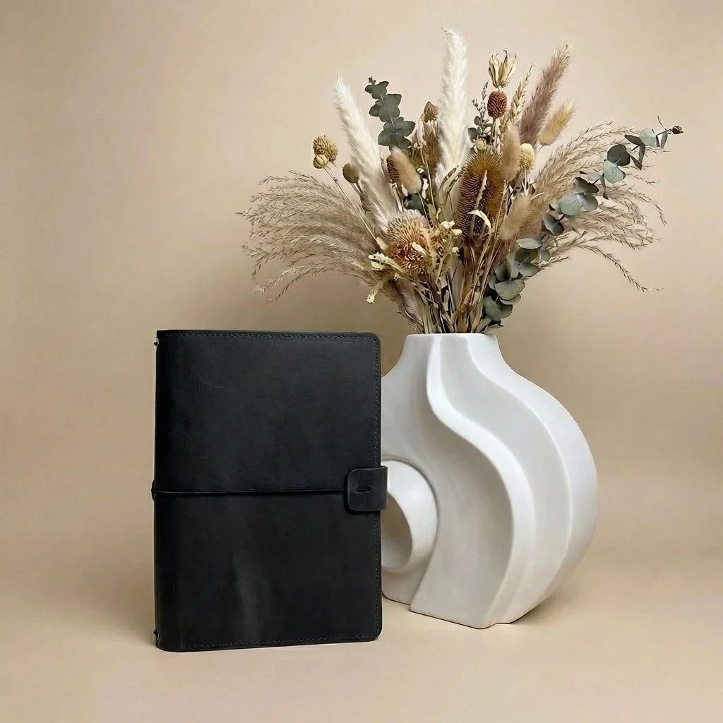 Black planner next to a white vase with dried plants on a beige background