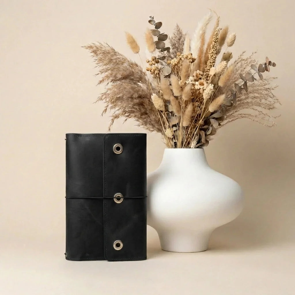 Black leather-bound notebook and white vase with dried plants on a beige background