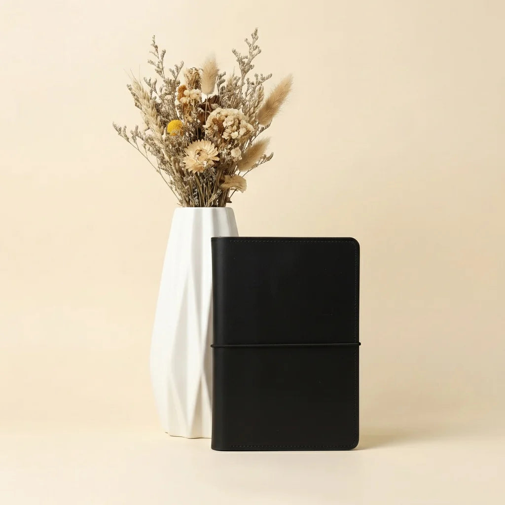 Black notebook next to a vase with dried flowers on a beige background