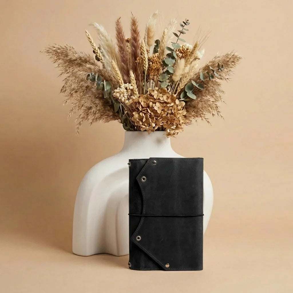 Black leather notebook with a white vase holding dried flowers on a beige background