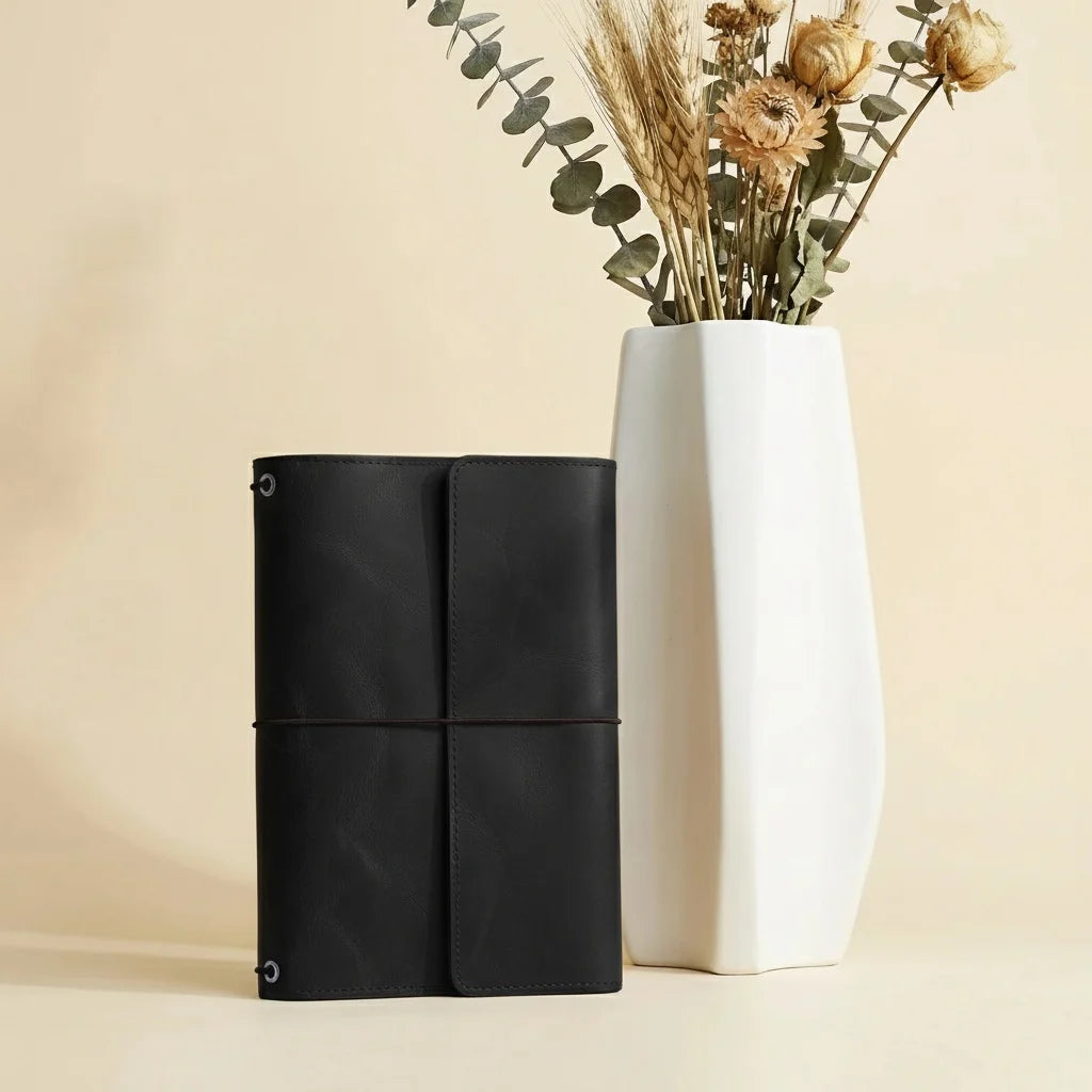 Black leather-bound notebook and white ceramic vase with dried flowers on a beige background
