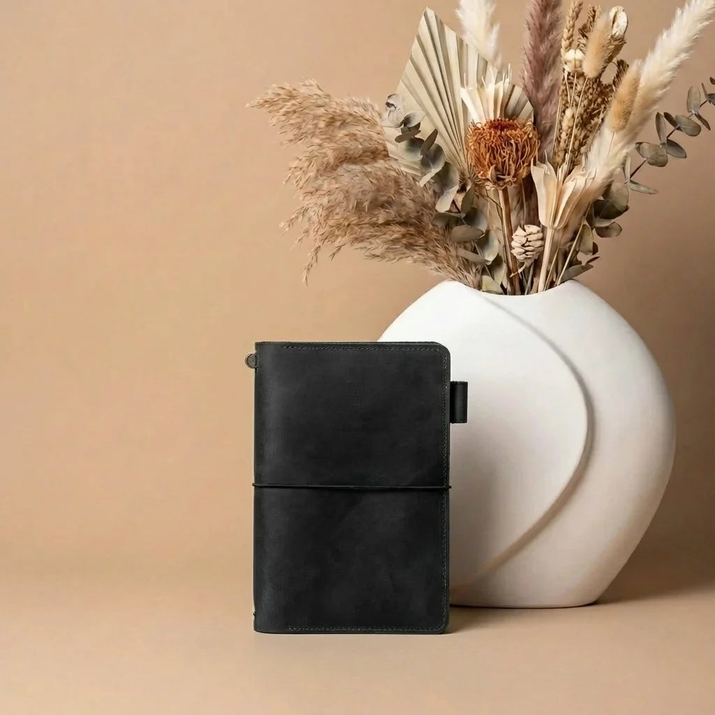 Black leather notebook next to a white vase with dried plants on a beige background