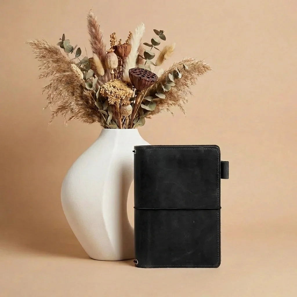 Black leather notebook next to a white vase with dried flowers on a beige background