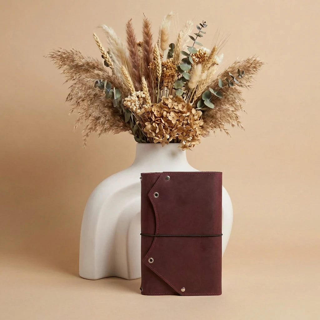 White vase with dried flowers and a brown leather notebook on a beige background