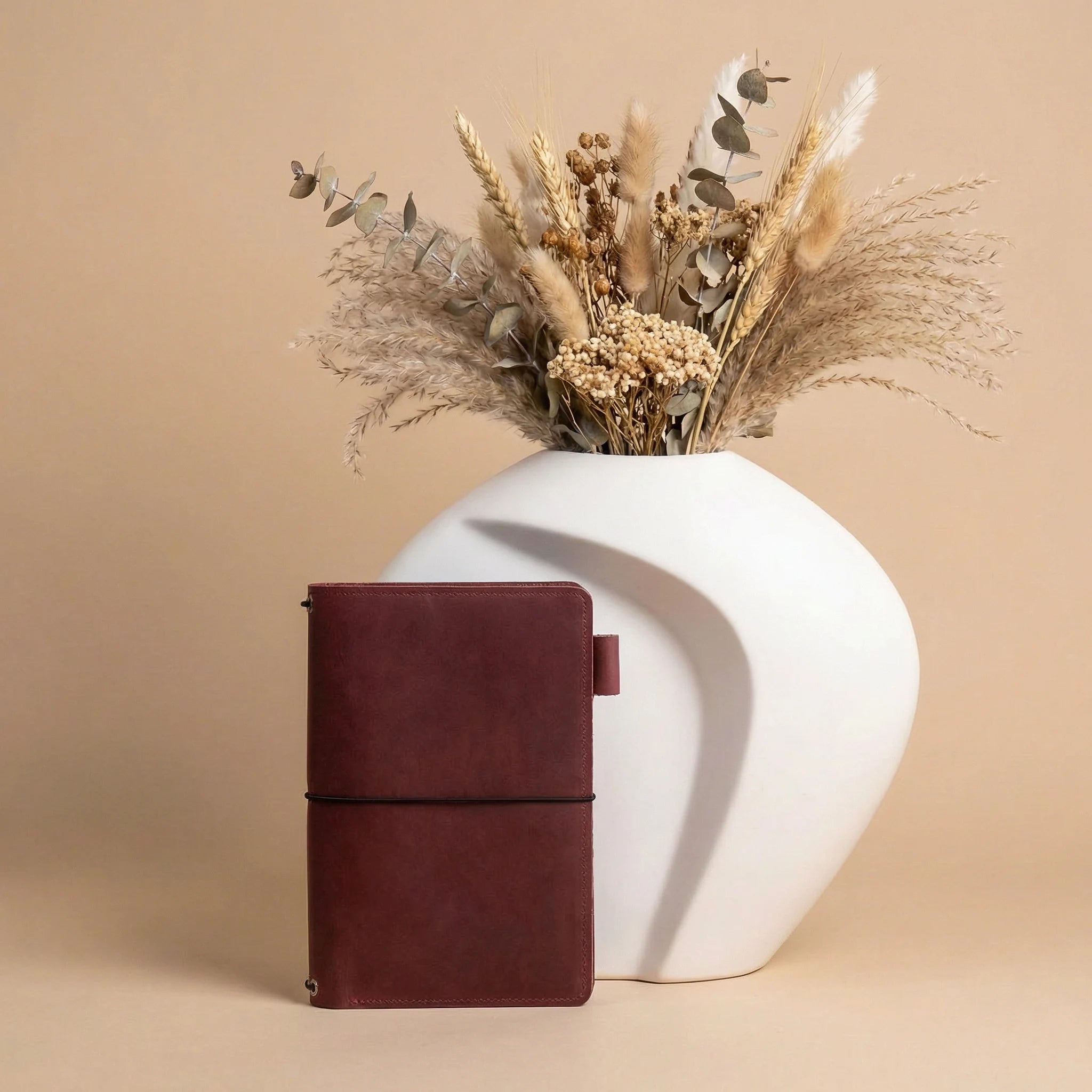 Brown leather notebook next to a white vase with dried flowers on a beige background
