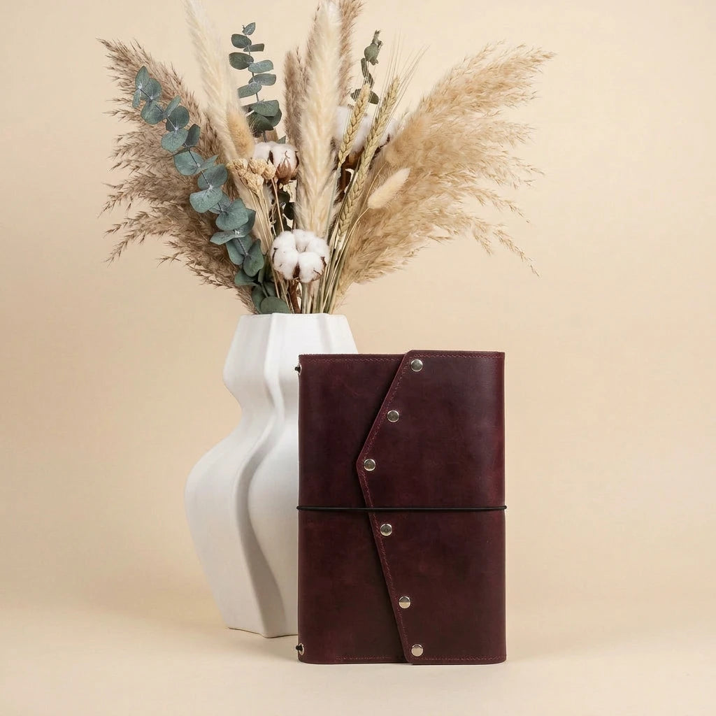 Brown leather notebook with silver buttons next to a white vase with dried plants on a beige background