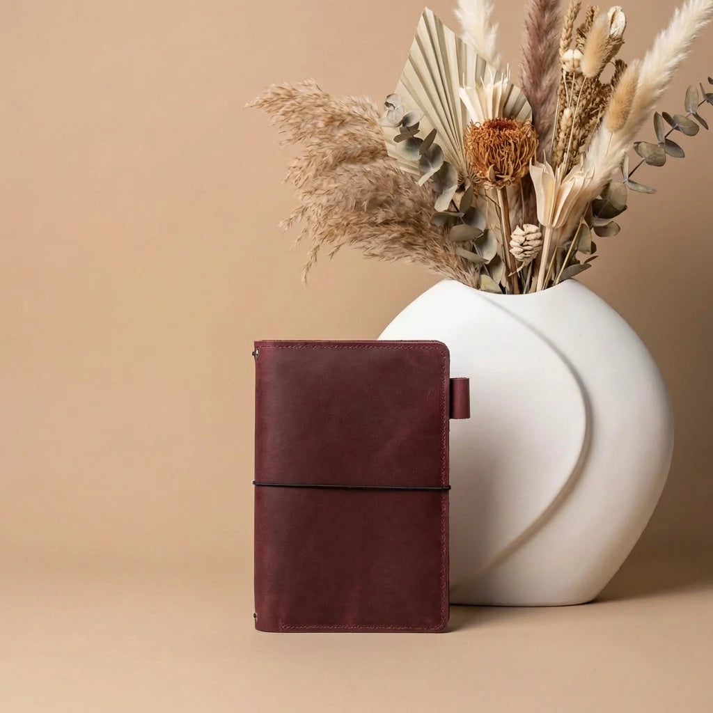 Brown leather notebook next to a white vase with dried plants on a beige background