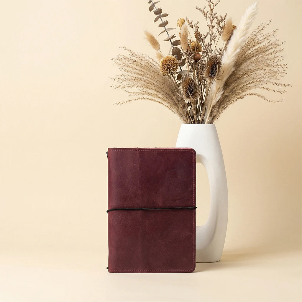 Brown leather notebook next to a white vase with dried plants on a beige background