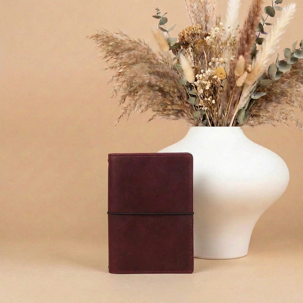 Brown leather notebook next to a white vase with dried flowers on a beige background