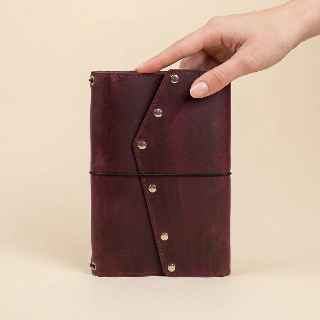 Hand holding a burgundy leather notebook with metal rivets on a beige background