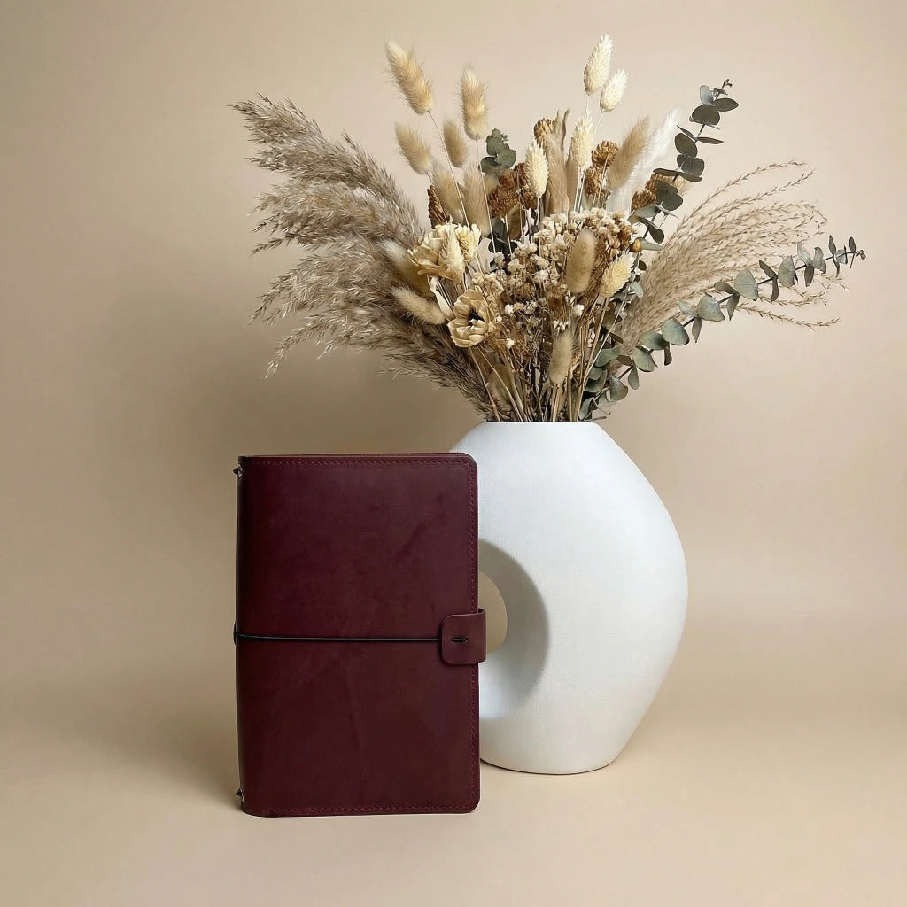 Brown leather notebook next to a white vase with dried flowers on a beige background