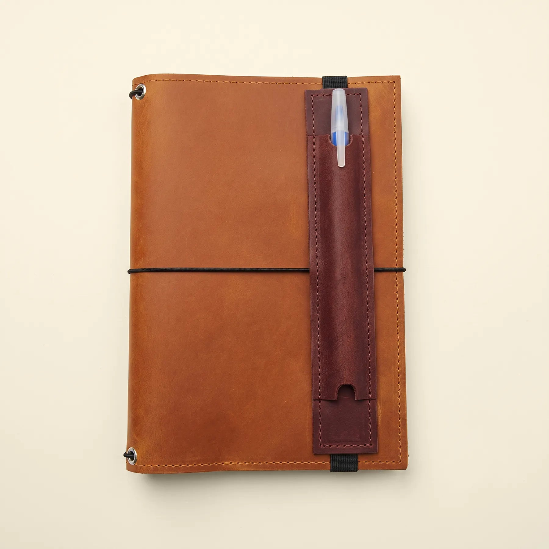 Compact leather pen holder attached to notebook cover with flexible elastic strap.