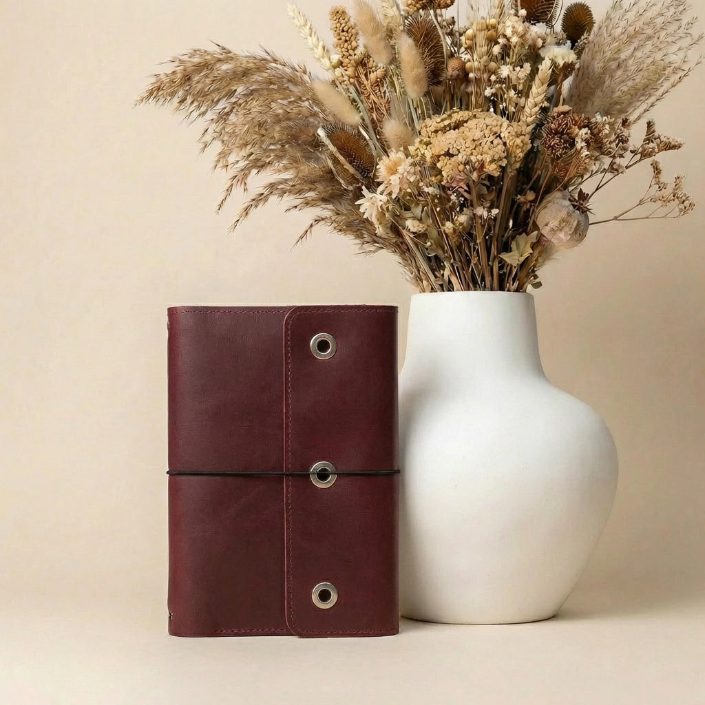 Brown leather notebook next to a white vase with dried flowers on a beige background