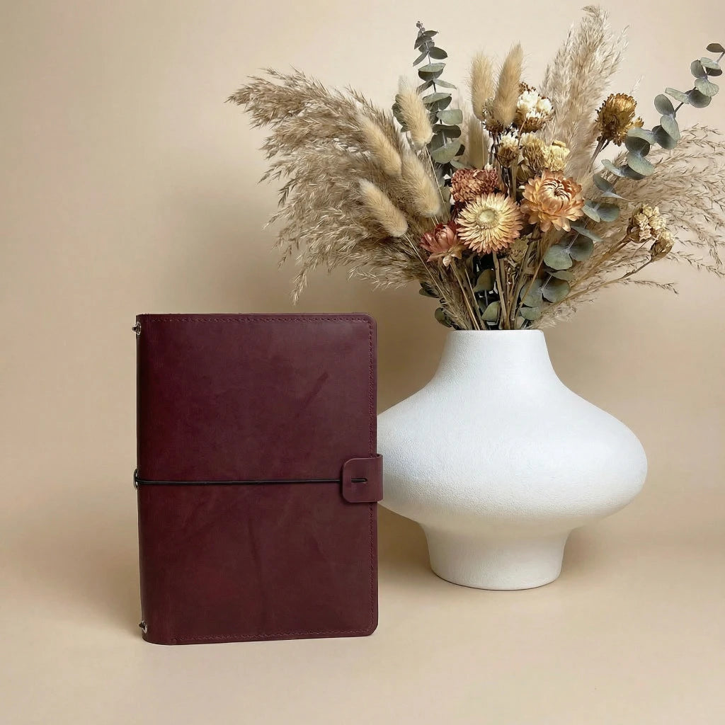 Brown leather notebook next to a white vase with dried flowers on a beige background