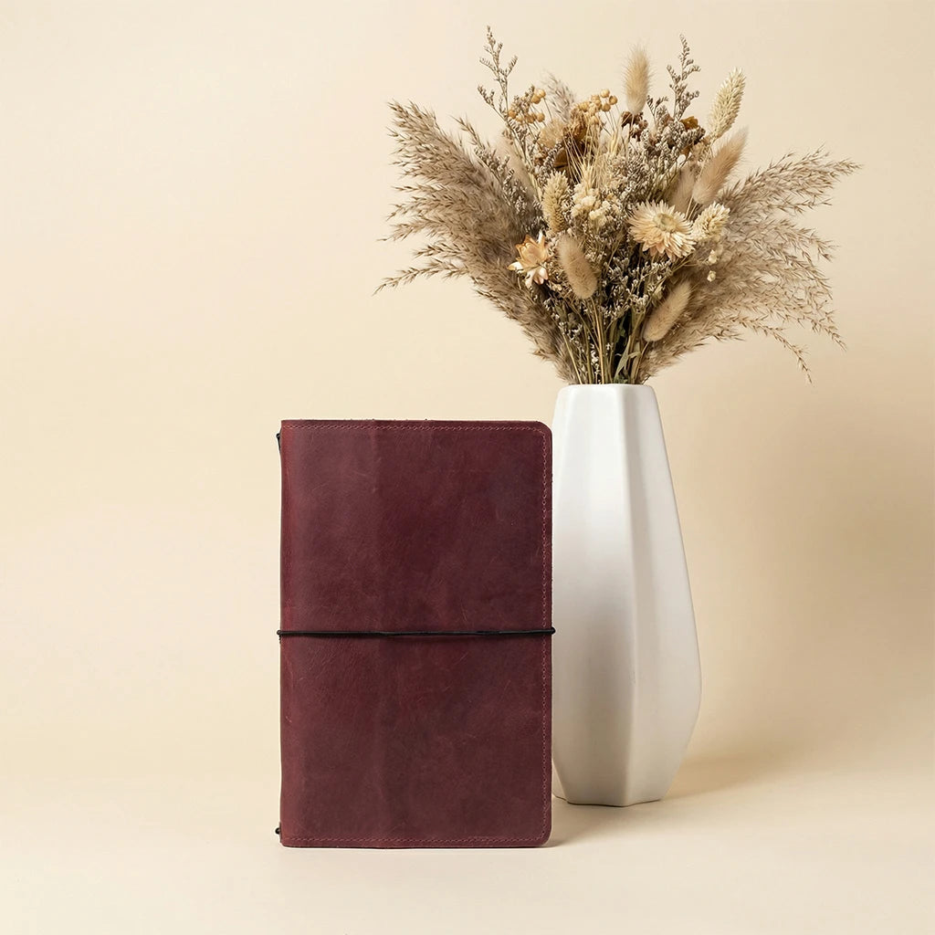 Maroon leather notebook next to a white vase with dried flowers on a beige background