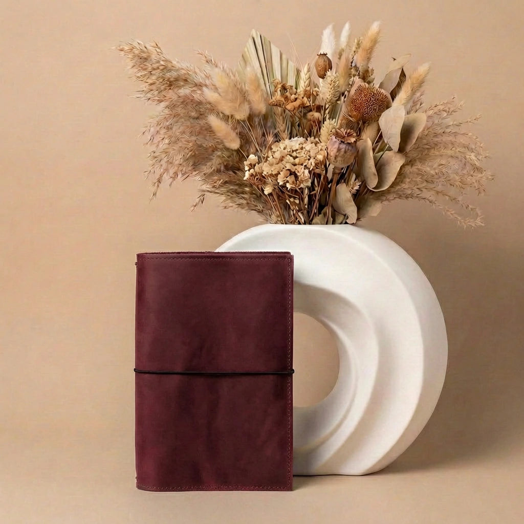Brown leather notebook next to a white vase with dried flowers on a beige background