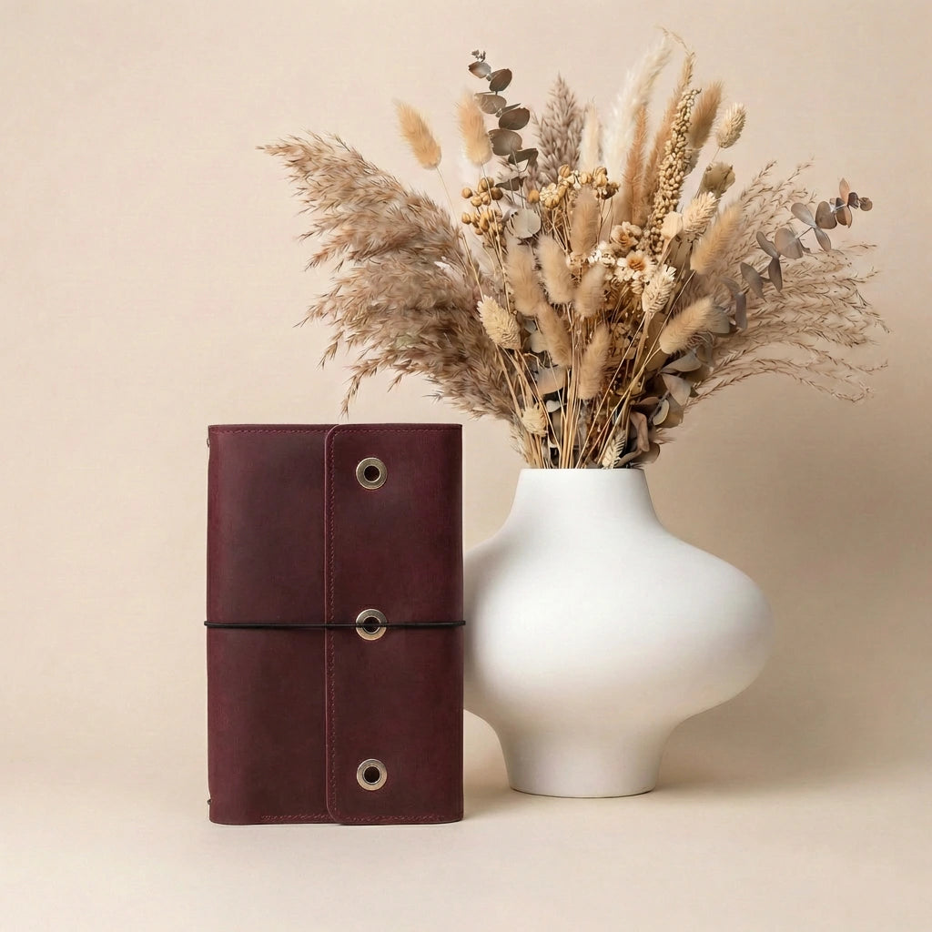 Brown leather notebook next to a white vase with dried flowers on a beige background