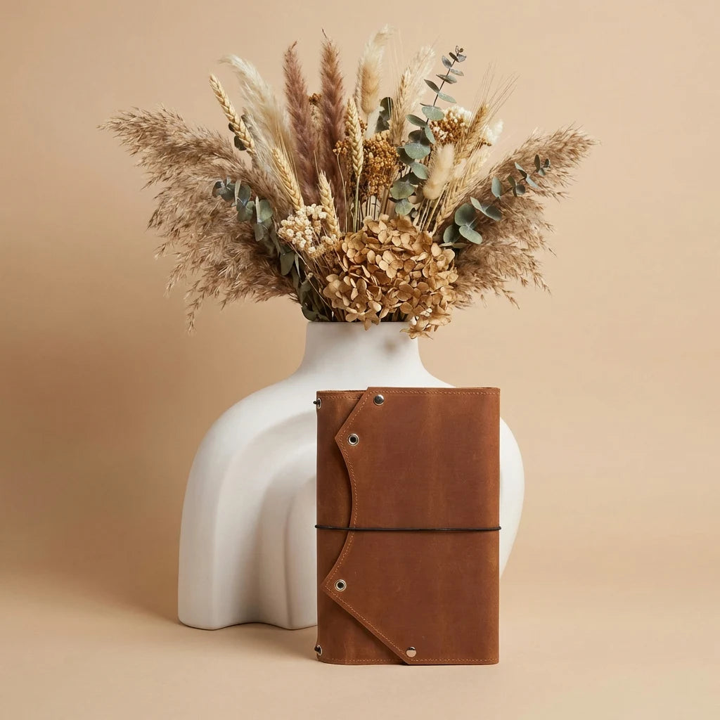 White vase with dried flowers and a brown leather notebook on a beige background
