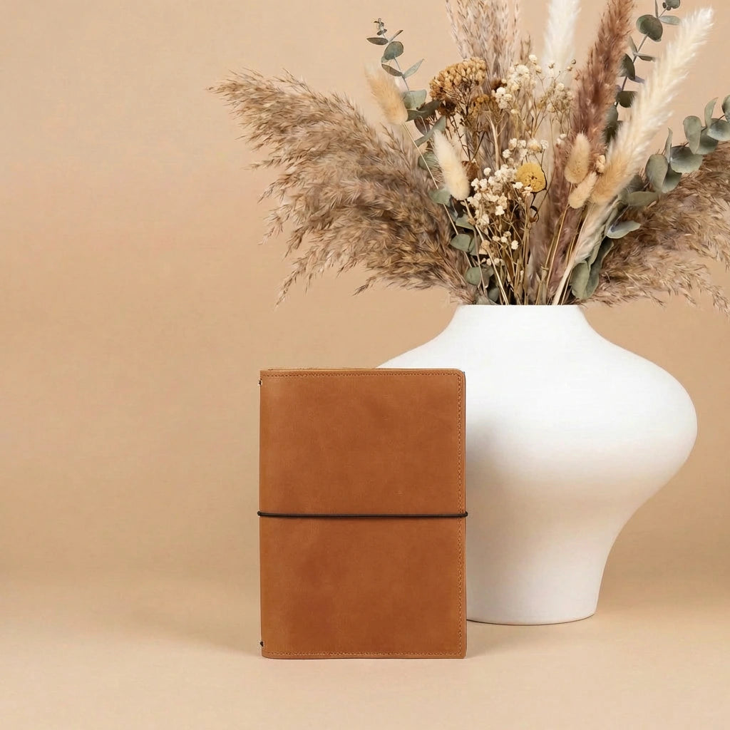 Brown leather notebook next to a white vase with dried flowers on a beige background