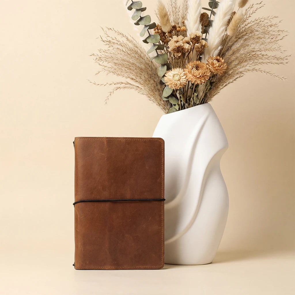 Brown leather notebook next to a white vase with dried flowers on a beige background