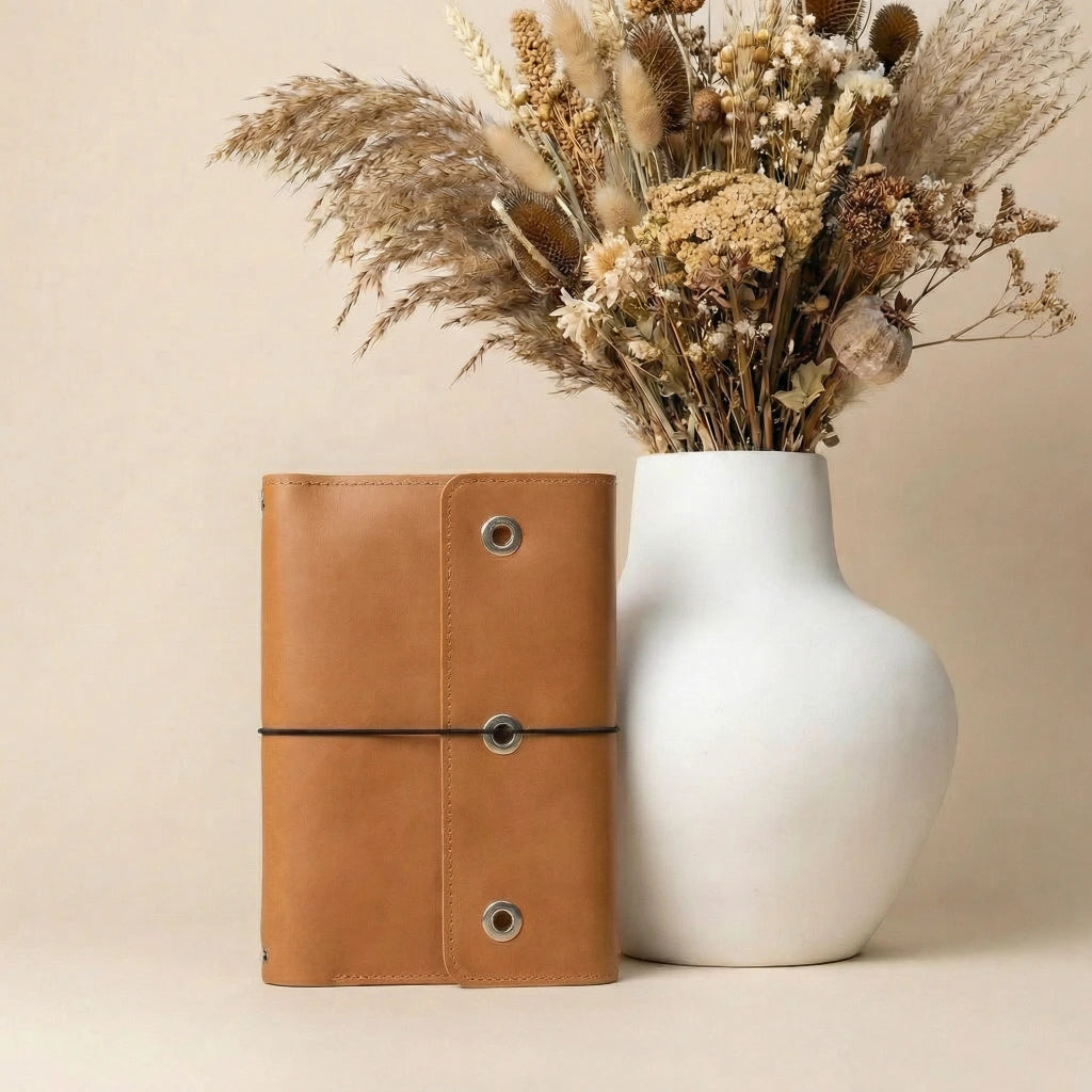 Brown leather notebook and white vase with dried flowers on a beige background