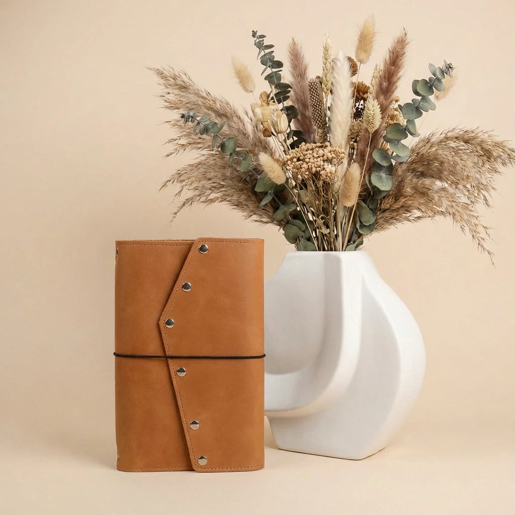Brown leather journal next to a white vase with dried flowers on a beige background