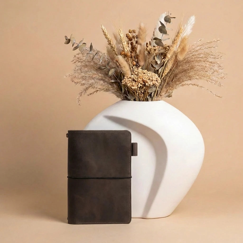 Black leather notebook next to a white vase with dried flowers on a beige background