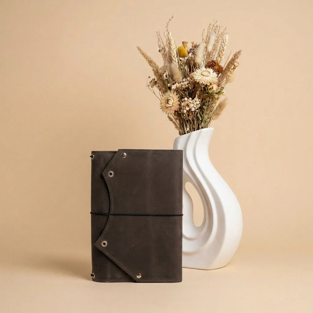 Brown leather notebook and white vase with dried flowers on a beige background