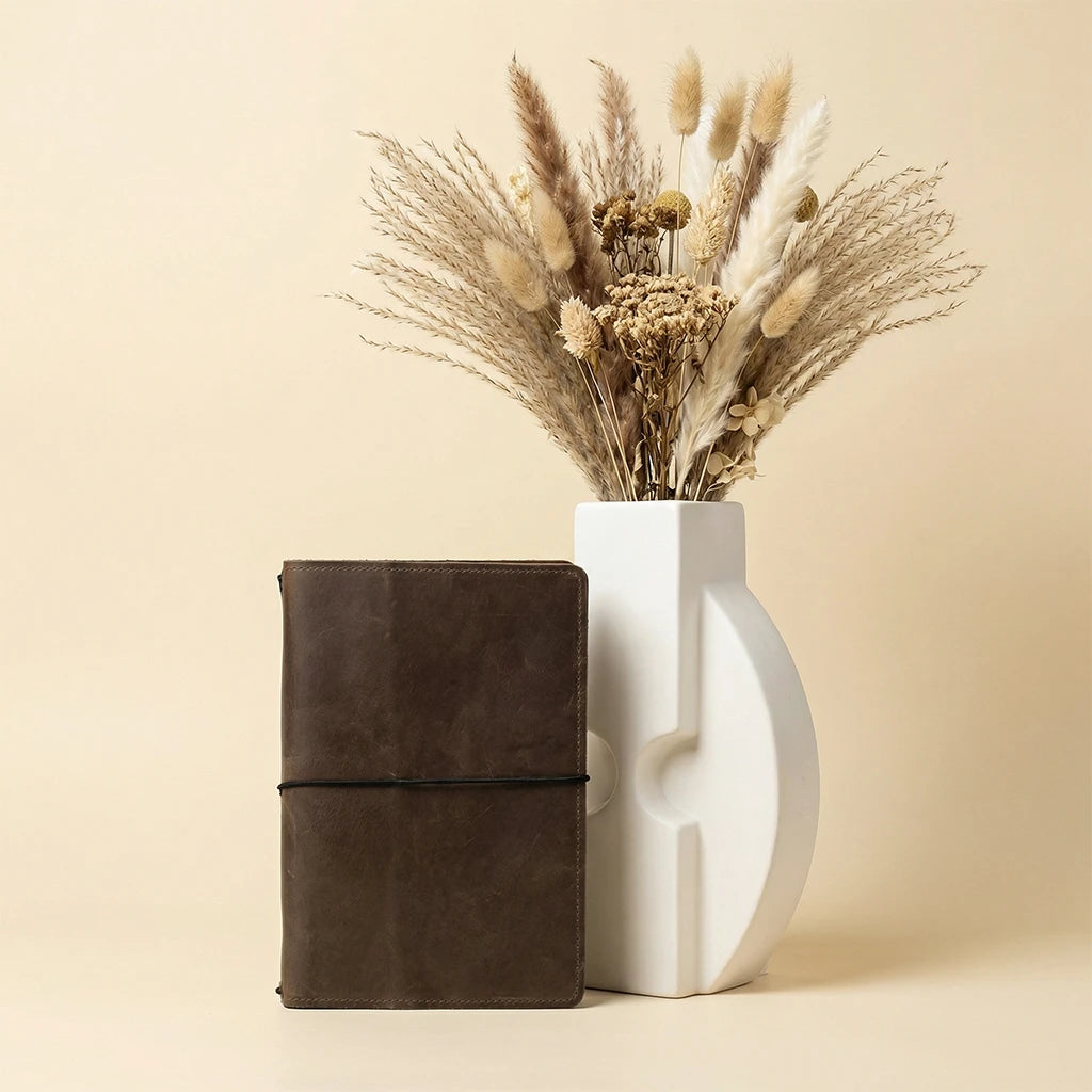 Brown leather notebook next to a white vase with dried plants on a beige background