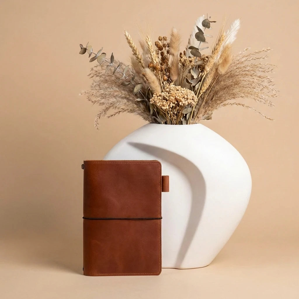 Brown leather notebook next to a white vase with dried flowers on a beige background
