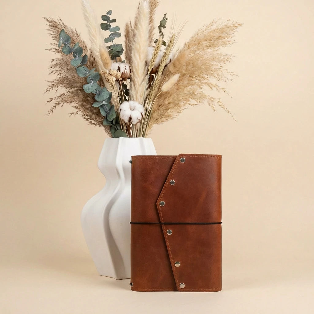 Brown leather notebook with decorative elements in front of a white vase with dried plants on a beige background