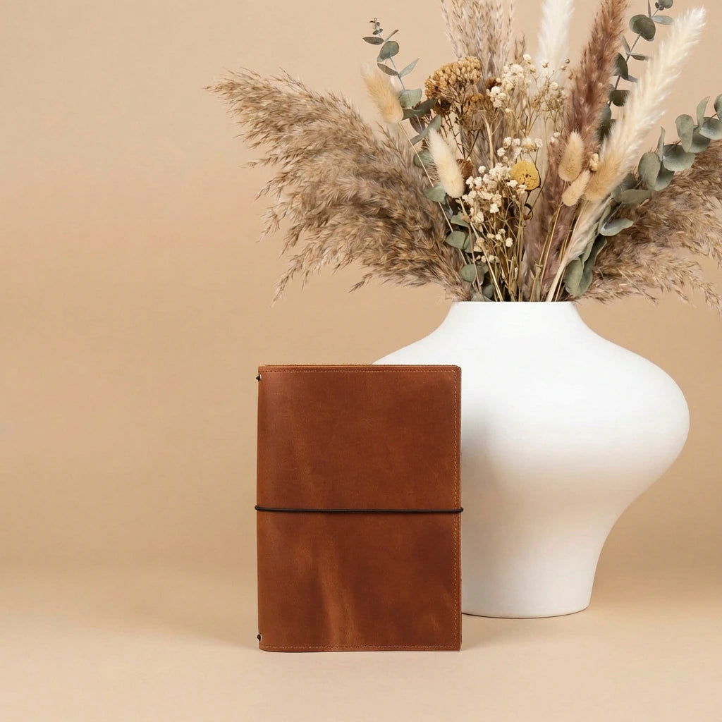 Brown leather notebook next to a white vase with dried flowers on a beige background