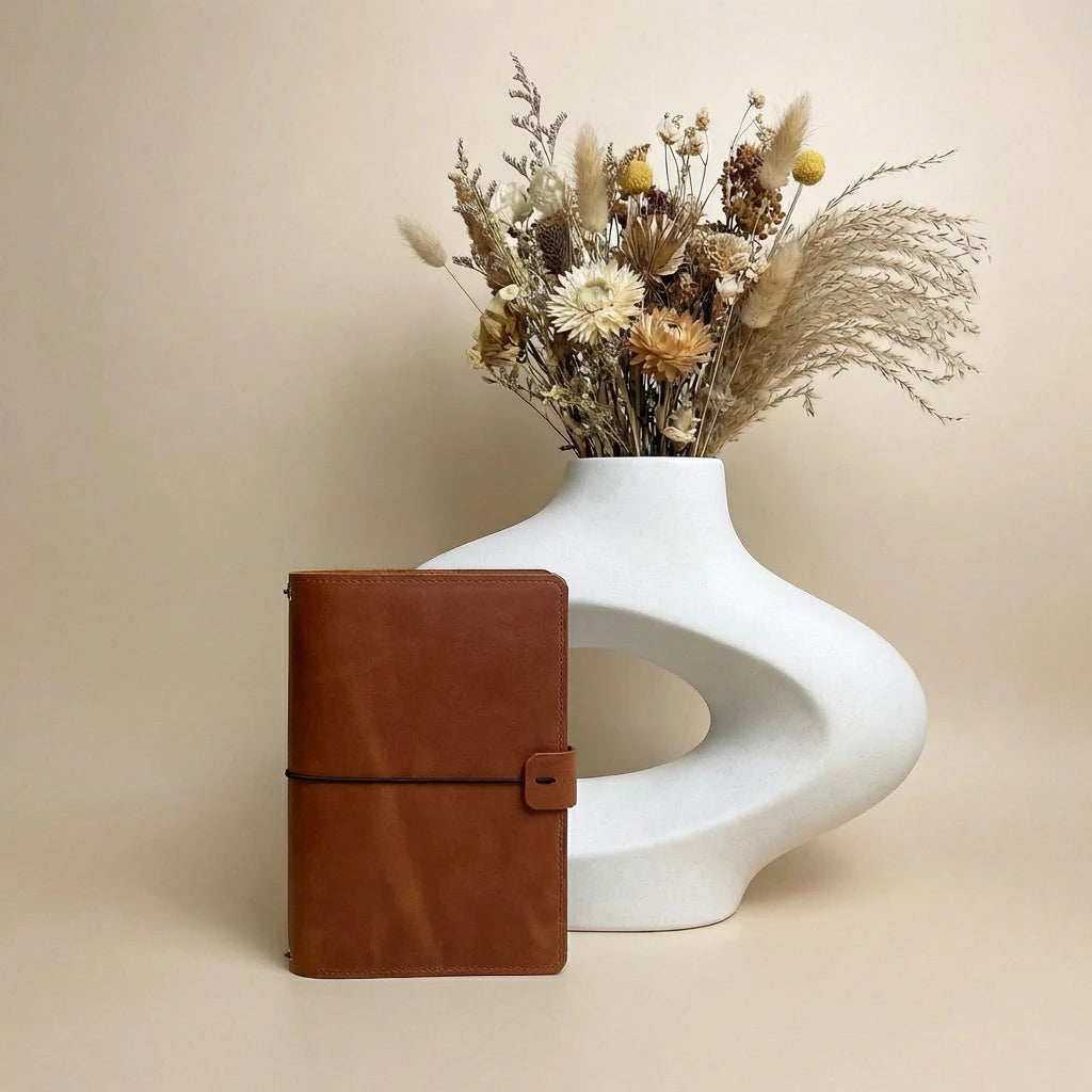 Brown leather notebook next to a white vase with dried flowers on a beige background