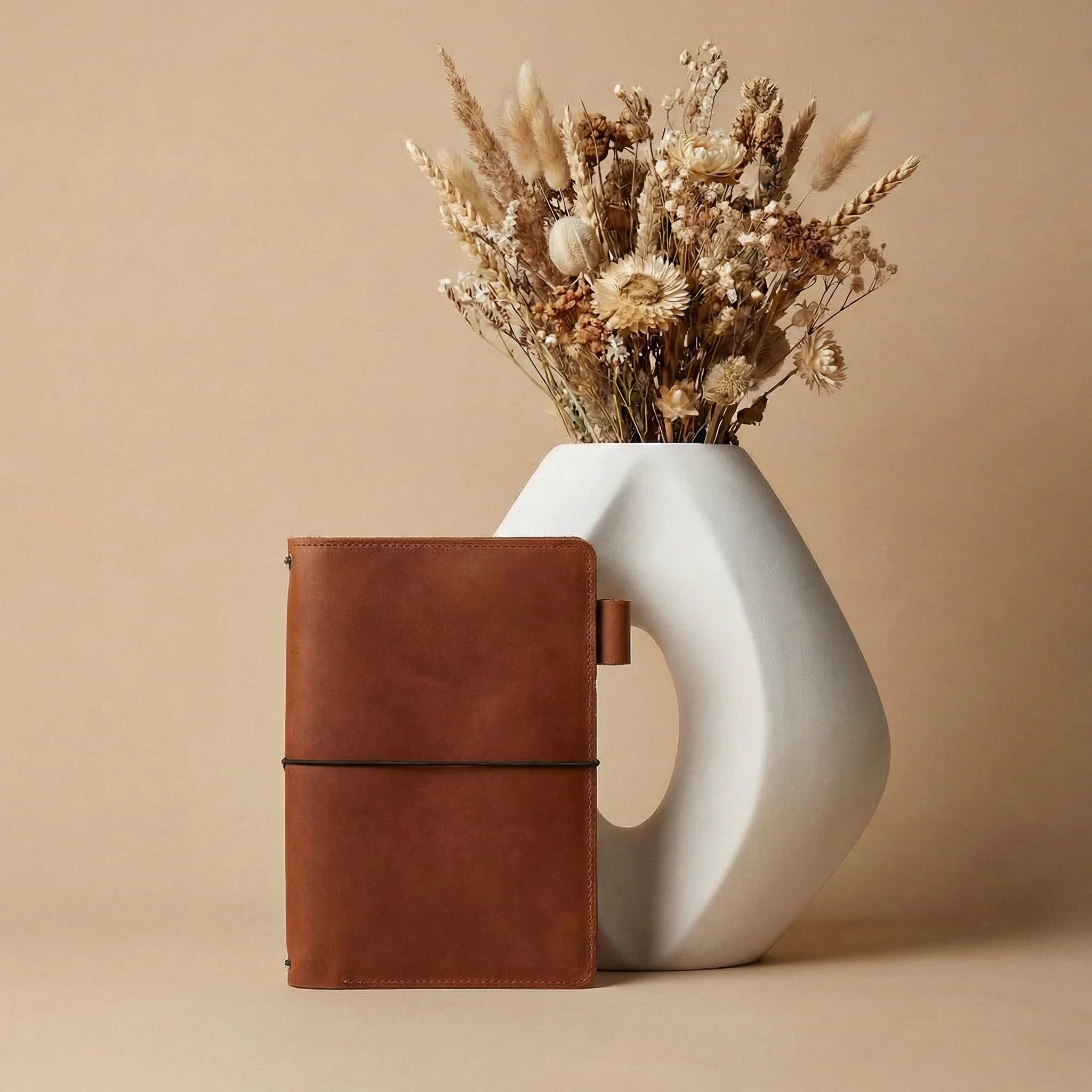 Brown leather notebook next to a white vase with dried flowers on a beige background