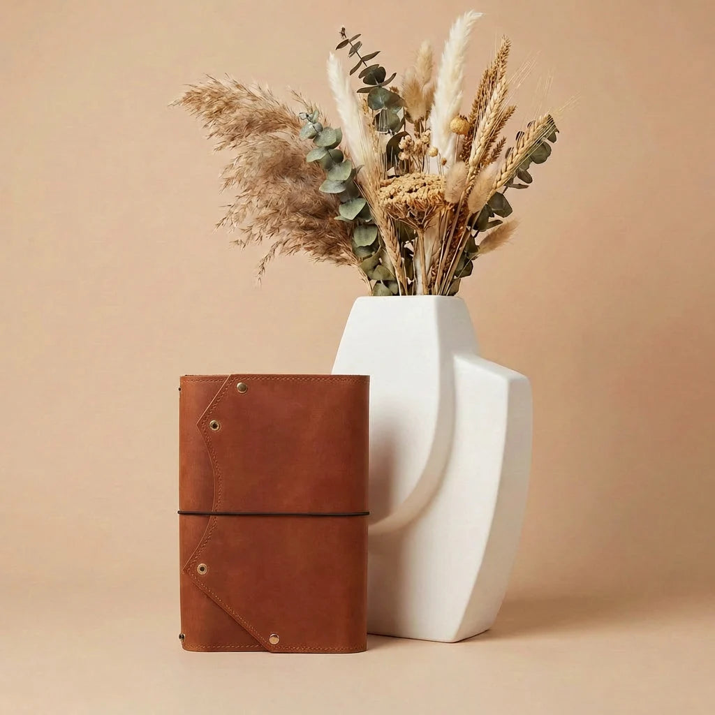 Brown leather notebook and white vase with dried plants on a beige background