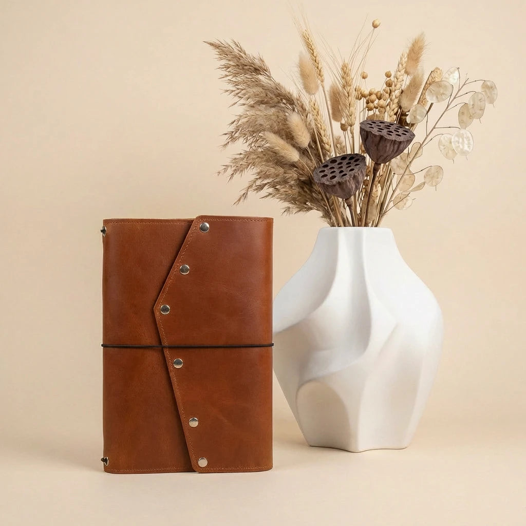 Brown leather notebook with silver buttons next to a white vase with dried flowers on a beige background
