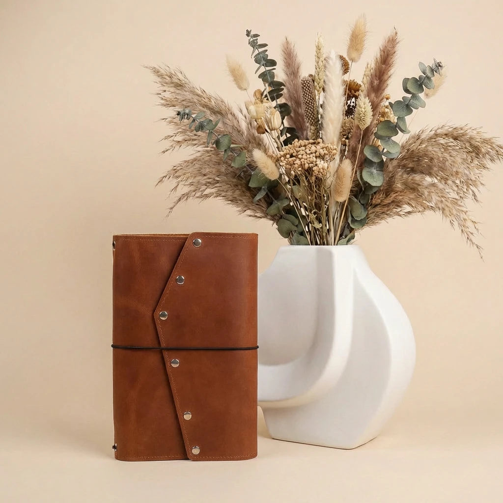 Brown leather notebook next to a white vase with dried flowers on a beige background
