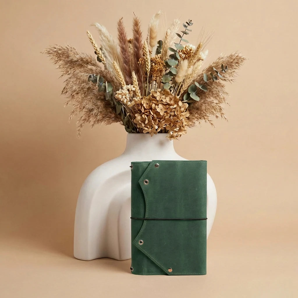 Green leather notebook in front of a white vase with dried flowers on a beige background
