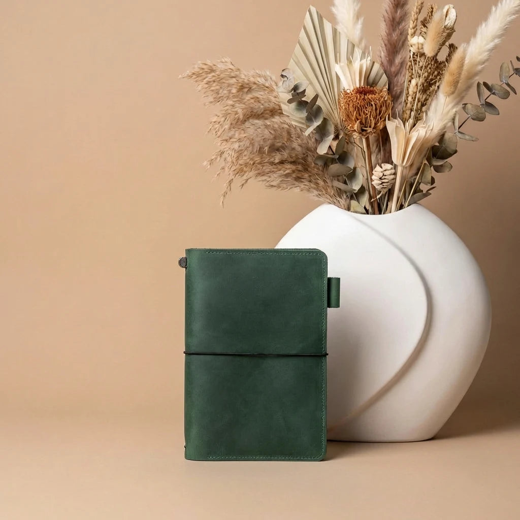 Green leather notebook next to a white vase with dried plants on a beige background