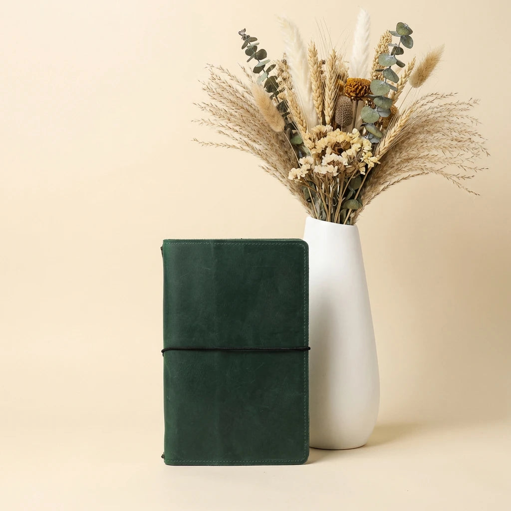 Green leather notebook next to a white vase with dried flowers on a beige background