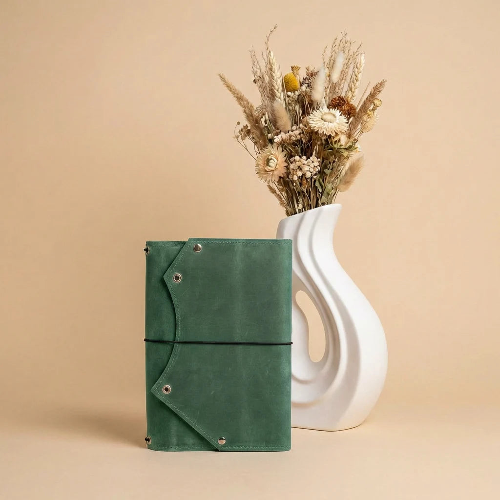 Green leather notebook next to a white vase with dried flowers on a beige background