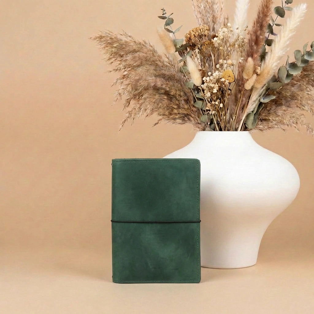 Green leather notebook next to a white vase with dried flowers on a beige background