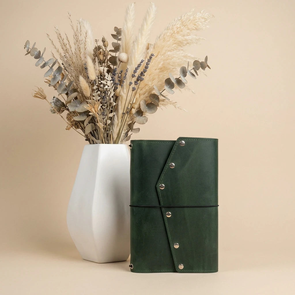 Green leather notebook with silver buttons next to a white vase with dried plants on a beige background