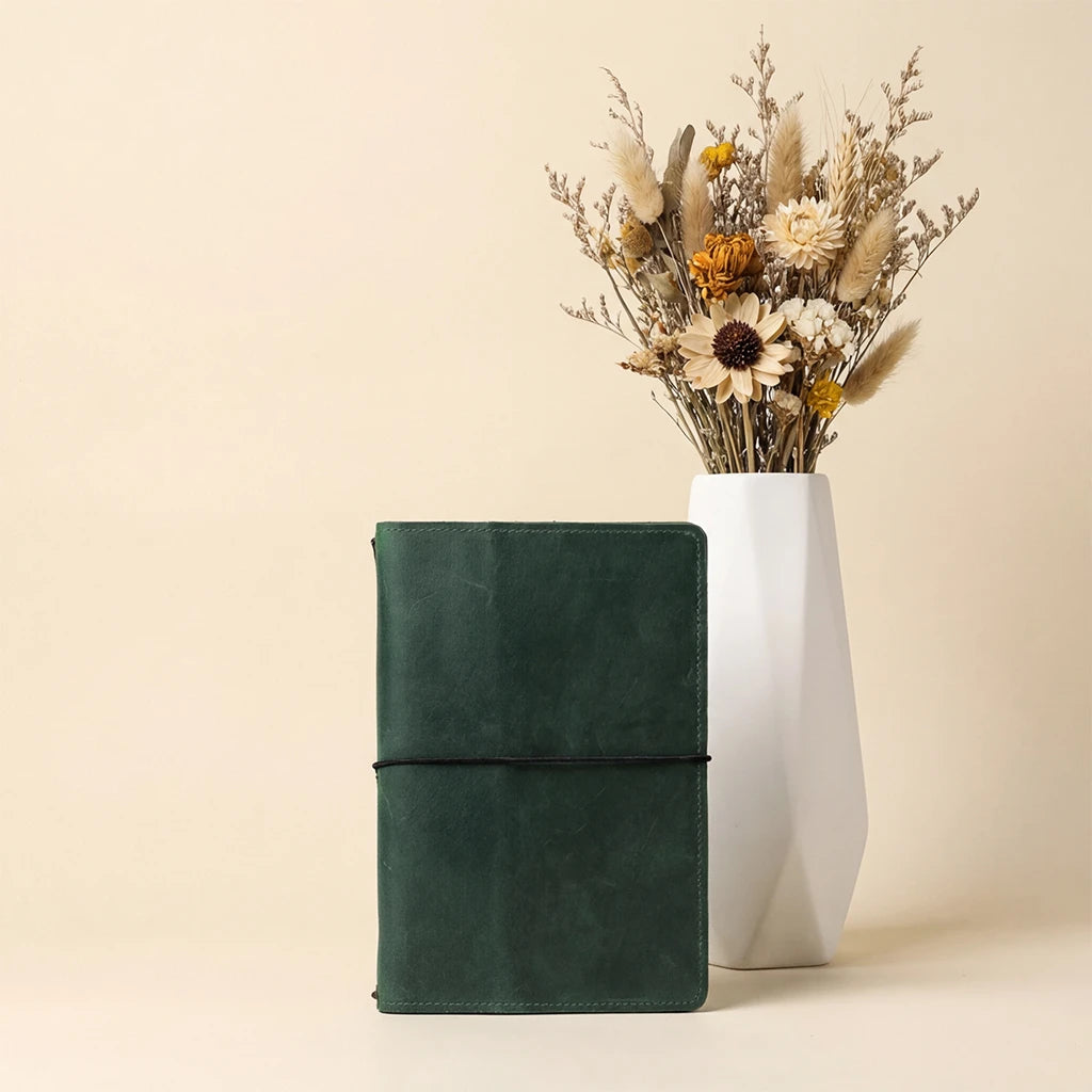 Green notebook next to a white vase with dried flowers on a beige background