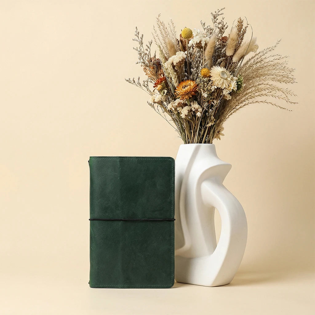 Green leather journal next to a white vase with dried flowers on a beige background