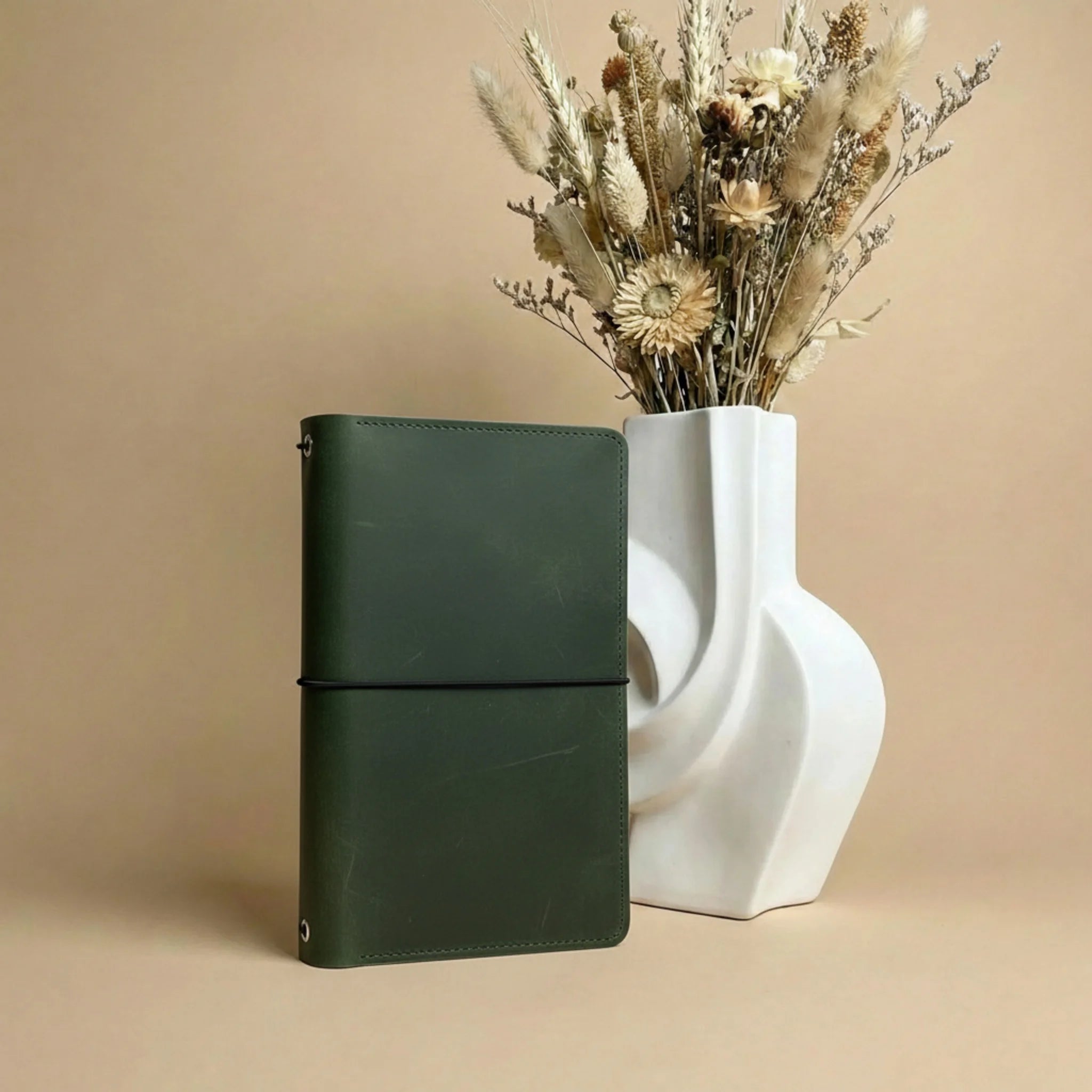 Green leather notebook next to a white vase with dried flowers on a beige background