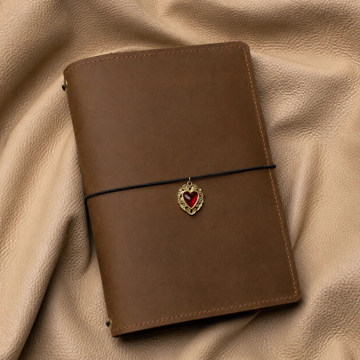 Decorative heart bead journal charm with metal clasp attached to refillable leather notebook elastic.