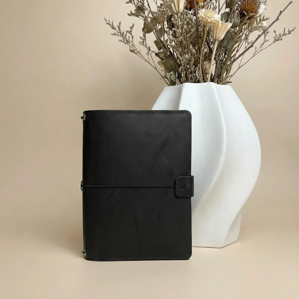 Black leather notebook next to a white vase with dried flowers on a beige background
